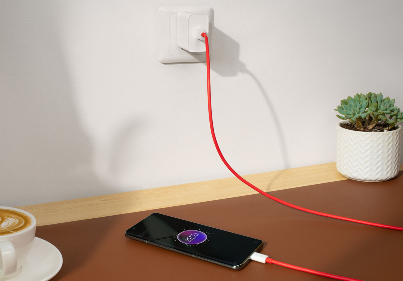 Charging a black smartphone with a red cable on a wooden table next to a succulent plant.