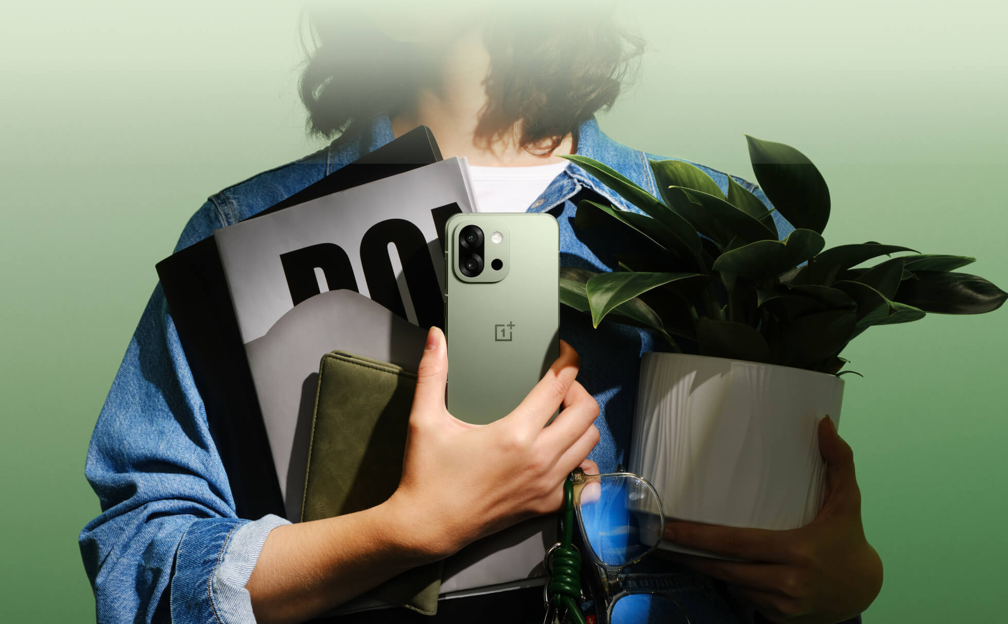 Человек в синей джинсовой куртке держит серый смартфон OnePlus и зеленое растение.